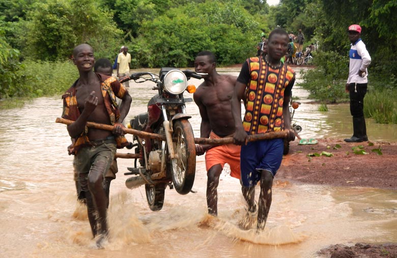 Das Bild zeigt eine mit braunem Wasser überflutete Straße zwischen grünen Büschen. Auf der Straße laufen vier junge Männer der Kamera entgegen, die  ein Motorrad auf zwei Holzstämmen tragen. Das Wasser reicht ihnen bis zu den Knöcheln und im Hintergrund beobachten einige Menschen die Aktion.