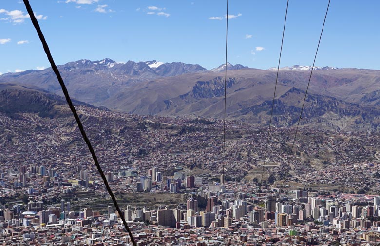 Eine Großstadt, die umgeben von Bergen größtenteils in einem Tal liegt, wird aus der Perspektive einer Seilbahngondel abgebildet. Im Vordergrund des Bildes: Seile und einzelne Gondeln.