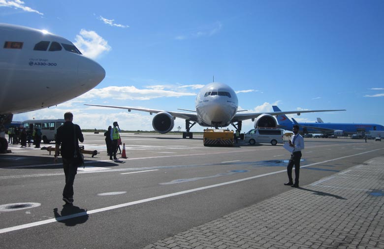 Drei Großraumflugzeuge auf einem Flugfeld, darüber ein strahlend blauer Himmel mit ein paar Wölkchen. Zwischen den Flugzeugen gehen ein paar Menschen und es stehen kleine Busse auf dem Flugfeld.