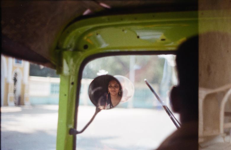 Das Bild wurde über die Schulter eines Tuk-Tuk-Fahrers fotografiert, so dass sein Hinterkopf noch unscharf und halb am rechten Bildrand zu sehen ist. Die Straße, die man durchs Fenster sieht, ist ebenso unscharf und der Fokus liegt auf  dem Seitenspiegel, in dem sich das Gesicht einer jungen Frau, die hinter ihm sitzt, spiegelt.