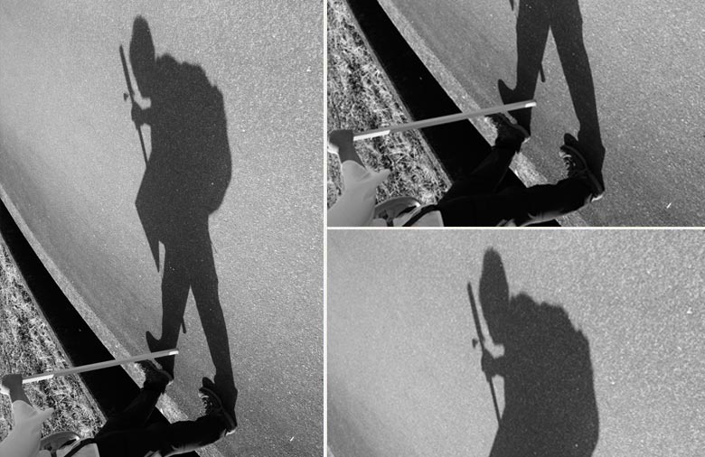 Das schwarzweiße Bild im Hochkantformat zeigt den Schatten einer Person mit Wanderstock und Rucksack, der sich auf einer Straße abzeichnet. Um dem Schatten Ausdruck zu verleihen wurde das Bild so gedreht, dass dieser aufrecht geht und die Person, die den Schatten wirft, ist bis auf die Beine und den Arm mit dem Wanderstock abgeschnitten.