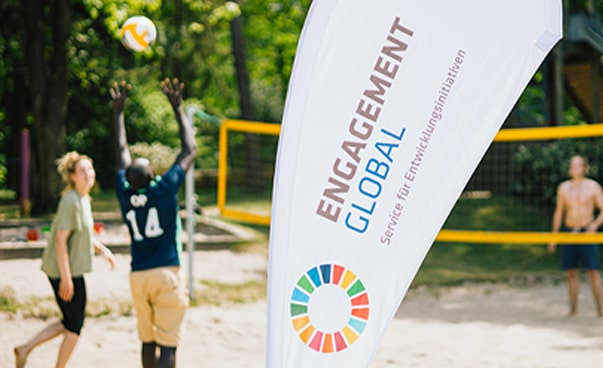 Eine Fahne mit der Aufschrift Engagement Global im Vordergrund. Im Hintergrund sind Teilnehmende, die Volleyball spielen.