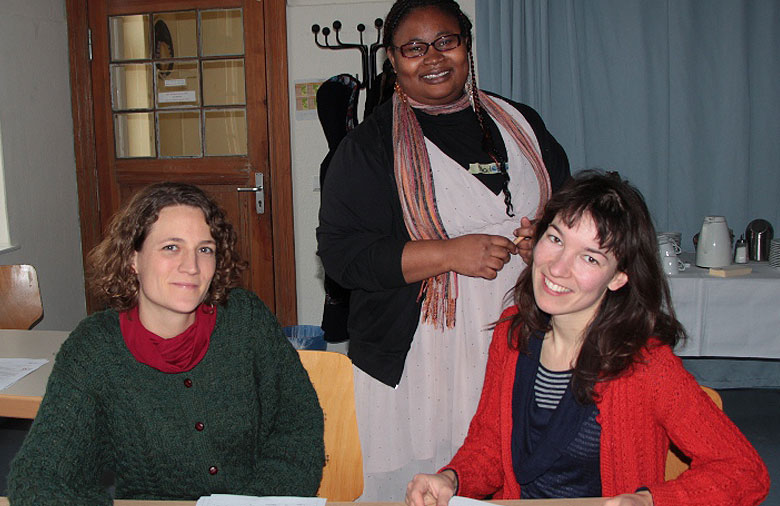 Zwei Personen sitzen an einem Tisch und lesen Bewerbungen. Hinter ihnen steht eine weitere Person. Alle drei lächeln in die Kamera. Foto: Miriam Eckert.