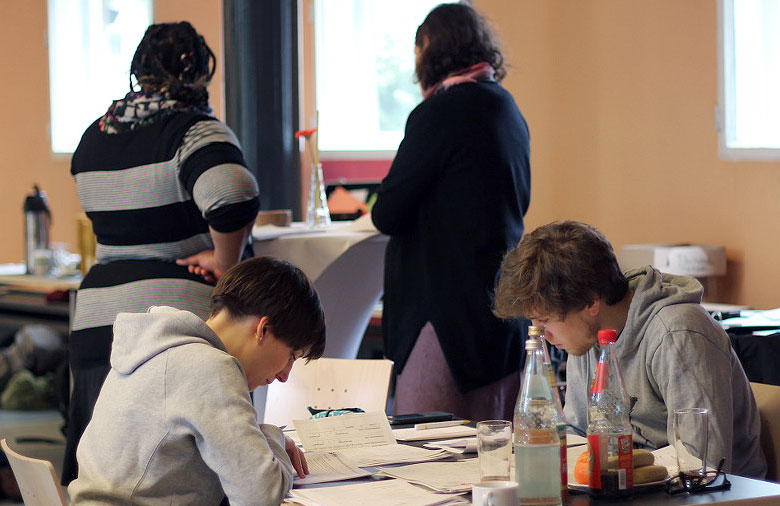 Zwei Personen sitzen an einem Tisch und lesen die Bewerbungen. Im Hintergrund stehen zwei weitere Personen mit dem Rücken zur Kamera. Foto: Mira Schönegge