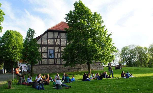 Ein Gutshaus steht auf einer grünen Wiese. Eine größere Gruppe junger Menschen sitzen und liegen auf dem Rasen davor unter Bäumen. Foto: Bernadette Lier