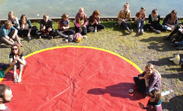 Mehrere Personen sitzen und stehen um eine runde, rote Plane auf dem Rasen.
