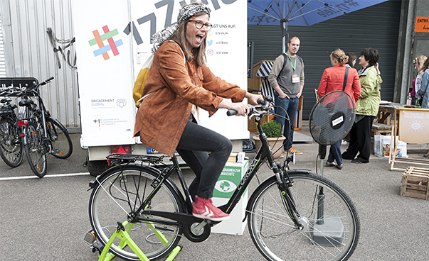 Junge Frau auf einem Fahrrad, Im Hintergrund unterhält sich eine Gruppe von Personen. Eine Leinwand mit #17Ziele steht hinter dem Fahrrad.
