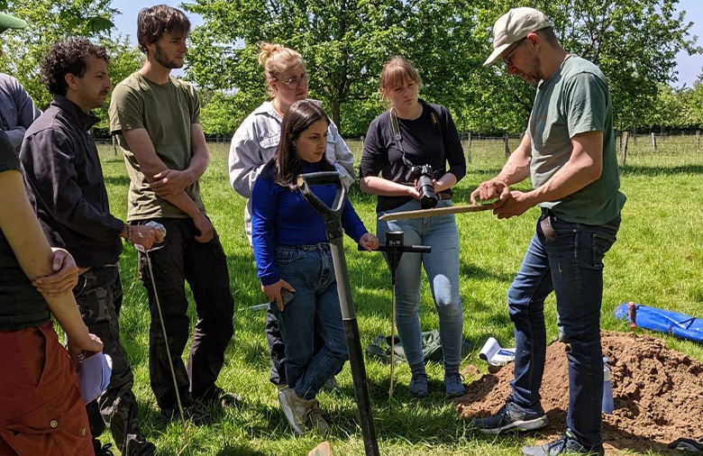 Students standing outdoors in front of a professor and a shovel.