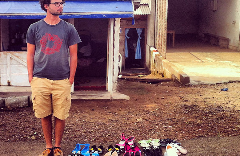 Ein junger Mann steht neben einer Reihe von verschiedenfarbigen Schuhen, die neben der Straße aufgereiht wurden. Im Hintergrund ist ein Haus oder eine Garage. Foto: Jonas Közle