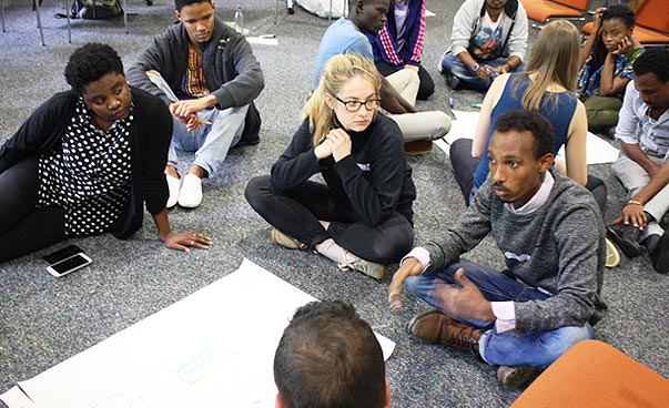 Junge Menschen auf dem Boden sitzend bilden zwei Kreise, zwischen ihnen liegt Flipchart-Papier. Sie scheinen miteinander zu diskutieren.