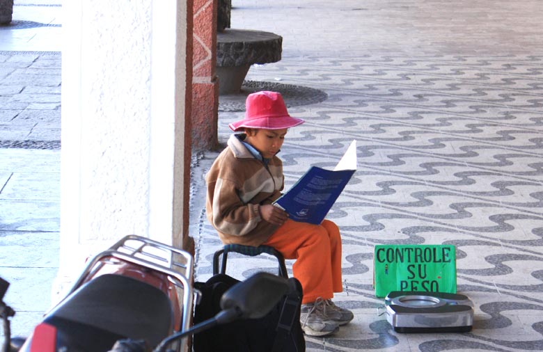 Ein kleiner Junge mit Hut sitzt auf einem kleinen Hocker in einer Fußgängerpassage und liest. Vor ihm ist ein grünes Schild aufgestellt, auf dem „Controle Su Peso“ geschrieben steht. Foto: Markus Wegenke