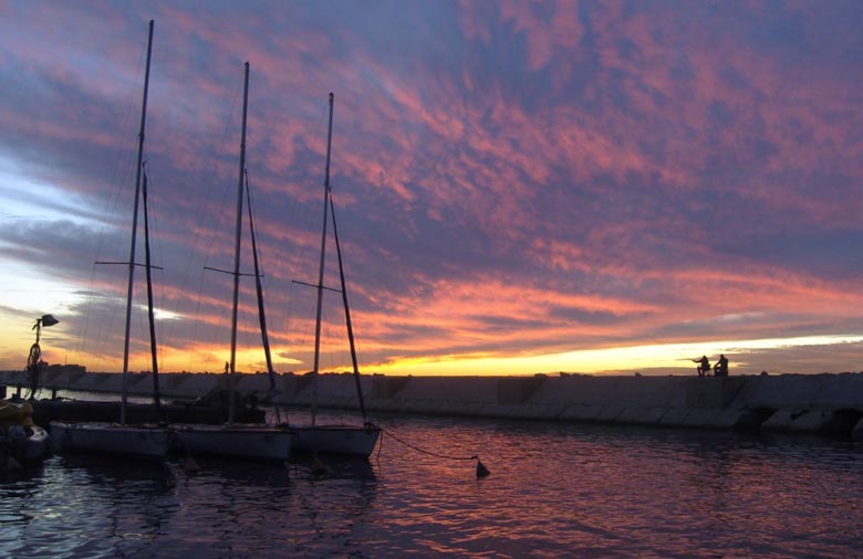 Ein Kanal oder das Meer bei Sonnenuntergang ist zu sehen. Auf der rechten Bildhälfte schwimmen Bote im Wasser. Links im Hintergrund zeichnen sich zwei Angler gegen den lila-rot-orange-gelb gefärbten Himmel ab. Foto: Hanna Blank