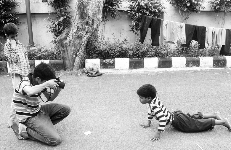 Drei Personen sind auf der schwarz-weiß Fotografie zu sehen. Ein Jugendlicher fotografiert einen Jungen, der sich auf die Straße gelegt hat und für die Kamera posiert. Ein anderer Junge steht neben dem Fotografen und beobachtet das Geschehen. Foto: Jens Marquardt