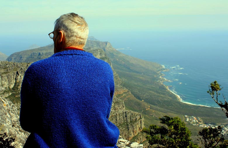 Von einem hohen Berg aus kann man das Meer und weitere Berge sehen. Ein Mann mit blauem Pullover sitzt mit dem Rücken zum Betrachter im Vordergrund auf einem Vorsprung. Foto: Antje Grebing.