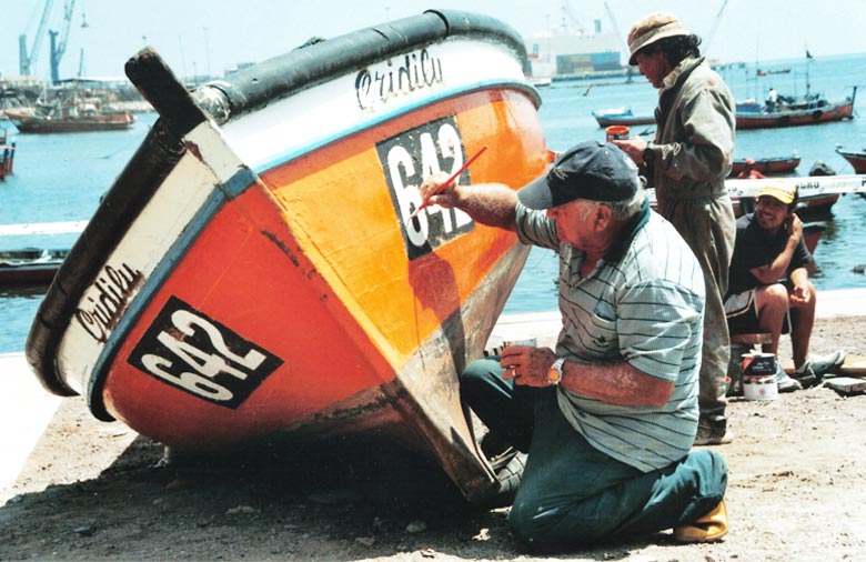 Ein Fischerboot wird von zwei Männern neu gestrichen. Im Hintergrund sieht man ein Hafenbecken. Foto: Torben Ibs