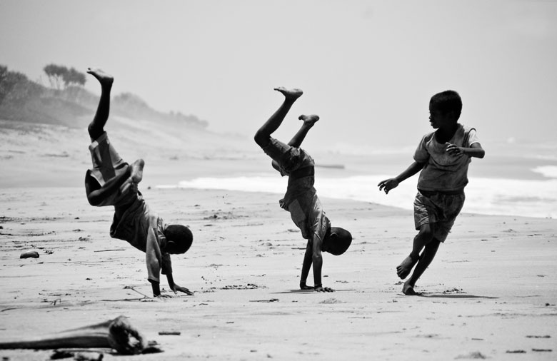 Das Bild ist in Schwarz-Weiß. Es ist ein Strand zu sehen auf dem zwei Jungs einen Radschlag machen; ein Junge rennt neben ihnen. Foto: Alina Wander, Simon Günzel.