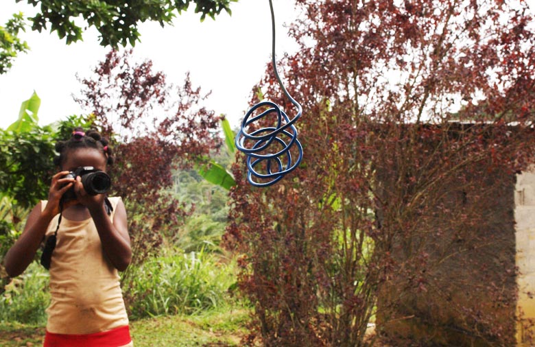 Ein Mädchen guckt durch eine Fotokamera. Sie fotografiert ein im Fokus hängendes Kabel. Im Hintergrund sind Palmen und andere Pflanzen. Foto: Caroline Erikson.