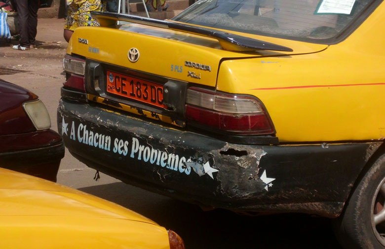 Der Bildausschnitt zeigt die Rückseite eines gelben Autos. Die Stoßstange ist stark abgenutzt und darauf steht in Sternen gerahmt geschrieben ‚A Chaqun ses Problèmes’ (‚Jedem seine Probleme’). Im Hintergund laufen Menschen.