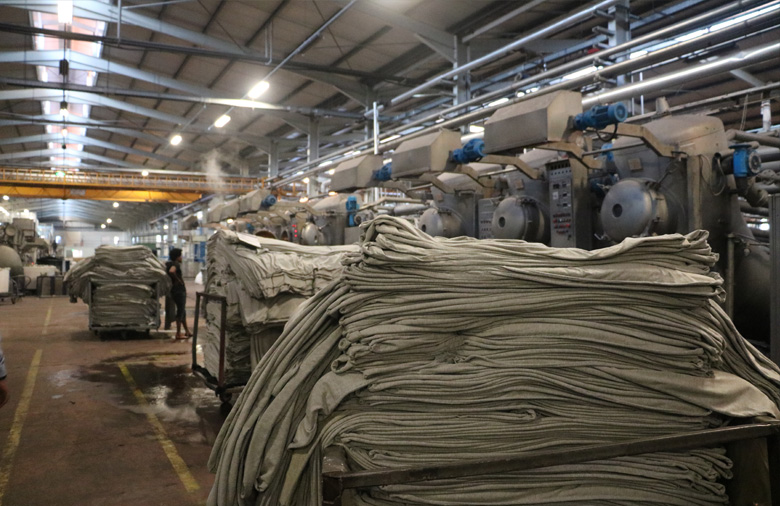 Innerhalb einer Fabrik stehen mit Stoffen vollgeladene Wägen vor großen Maschinen.