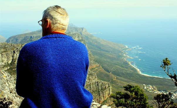 Von einem hohen Berg aus kann man das Meer und weitere Berge sehen. Ein Mann mit blauem Pullover sitzt mit dem Rücken zum Betrachter im Vordergrund auf einem Vorsprung. Foto: Antje Grebing