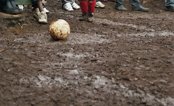Ein Fußball auf nasser Erde, im Hintergrund sind Füße zu sehen. Foto: Christina Seifert