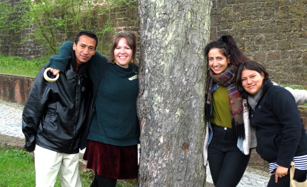 Vier junge Erwachsene stehen nebeneinander, die Arme um die Schulter der Nebenperson gelegt; in der Mitte ein Baumstamm. Foto: Celia Schmidt