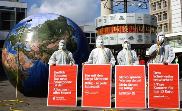 Vier Personen stehen in weißen Schutzanzügen stehen vor der Weltzeituhr auf dem Berliner Alexanderplatz und halten rote Plakate mit weißer Schrift. Hinter ihnen ist eine aufblasbare Skulptur einer Erdkugel. Foto: INKOTA