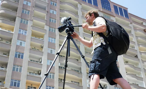 Eine Person steht mit einer Kamera, die auf einem Stativ befestigt ist, auf einer Mauer. Im Hintergrund ist ein Gebäude. Foto: Michael Kraus