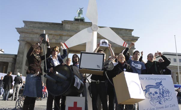 Eine Gruppe Menschen steht vor dem Brandenburger Tor und hat neben anderen Gegenständen ein Modell eines Windkraftwerks und eines Steckers in ihrer Mitte. Foto: Tim Gontrum
