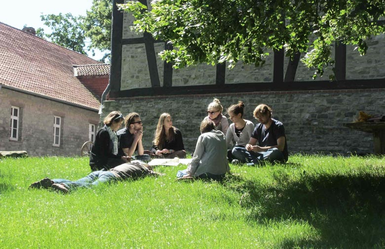 Eine Gruppe Menschen sitzt zusammen auf einem grünen Rasen. Ein Ast ragt ins Bild und im Hintergrund ist eine Fassade eines Fachwerkhauses zu sehen. Foto: Mira Schönegge
