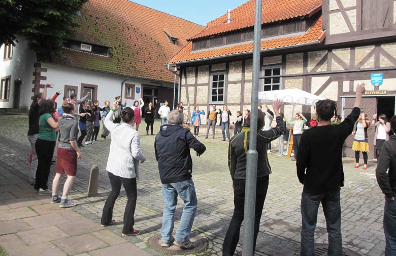 Eine große Gruppe Menschen steht zusammen in einem Kreis  und heben die Arme. Im Hintergrund sind Alte Fachwerkhäuser zu sehen. Foto: Mira Schönegge