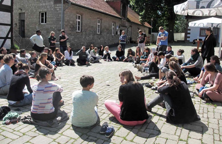 Eine große Gruppe Menschen sitzt  zusammen in einem Kreis im Freien. Im Hintergrund: alte Fachwerkhäuser. Foto: Mira Schönegge
