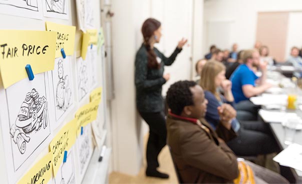 Das Bild zeigt ein Whiteboard mit Notizzetteln in Nahaufnahme. Im Hintergrund sind mehrere Personen, die sich in einer Unterrichtssituation befinden.