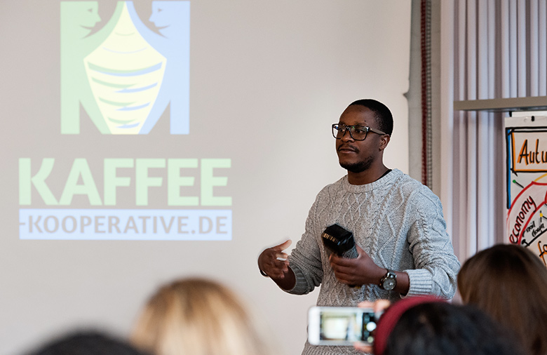 Allan Mubiru steht vor einer Leinwand, auf der „Kaffee-Kooperative.de“ steht. Er hat eine Packung schwarzen Kaffee in der Hand und gestikuliert mit seinen Händen. Im Vordergrund sind unscharf vier Köpfe von hinten zu sehen.