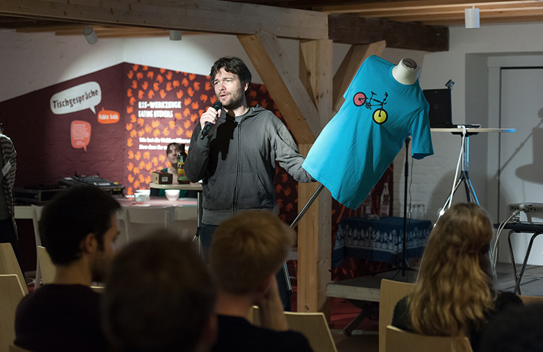 Stefan Niethammer von 3Freunde ist vor einem Publikum zu sehen. Er hat eine Kleiderpuppe mit einem blauen T-Shirt in der Hand und in der anderen hält er das Mikrophon.