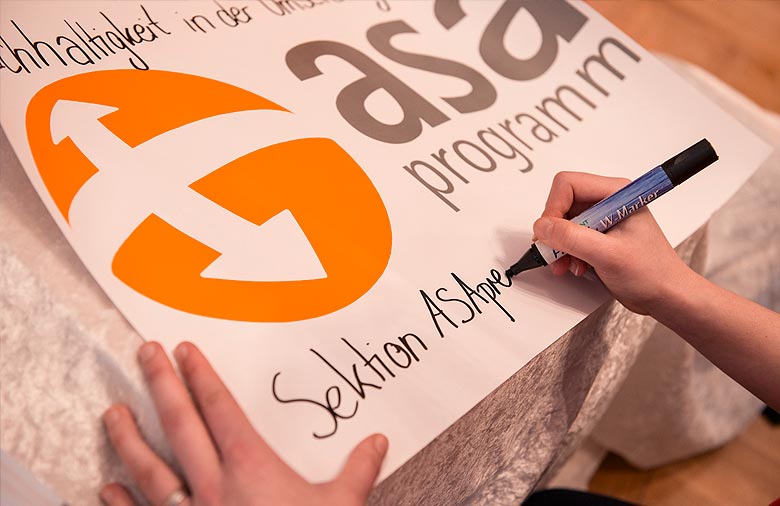 Eine Hand mit einem Stift schreibt „Sektion ASApreneurs“ auf ein Plakat mit dem Logo des ASA-Programms. Foto: Julia Kneuse.