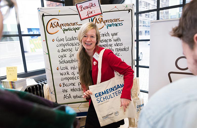 Natalie Campbell trägt eine Tasche mit der Aufschrift „Gemeinsam Schultern“. Sie steht vor einem Plakat, dass die Vorteile von ASApreneurs für Teilnehmende und Partner erläutert. Foto: Jule Roehr.